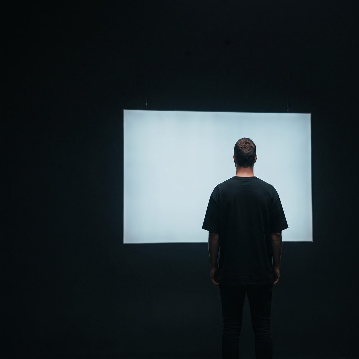 A man seen from behind, standing in a dark room facing a large blank illuminated screen.