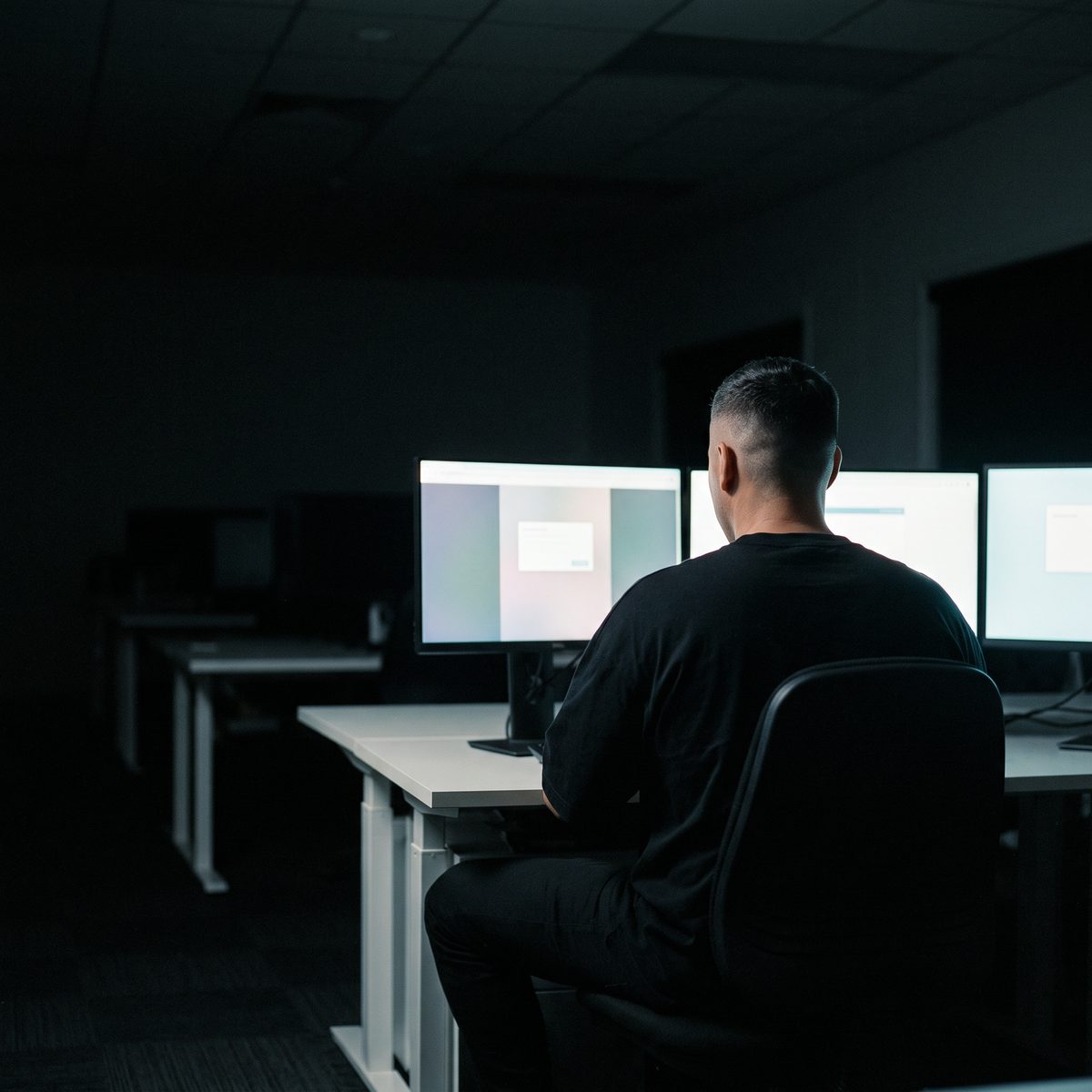 A man seen from behind, seated at a desk facing three glowing monitors in a dark office.