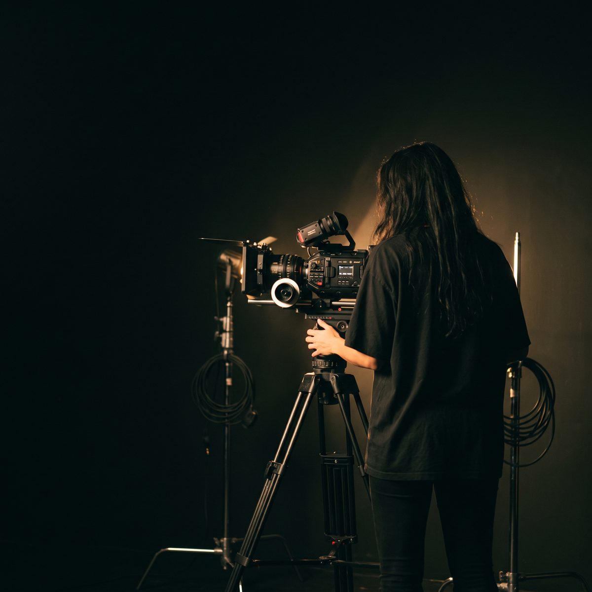 A woman with long dark hair, seen from behind, operating a cinema camera on a tripod in a dark studio.
